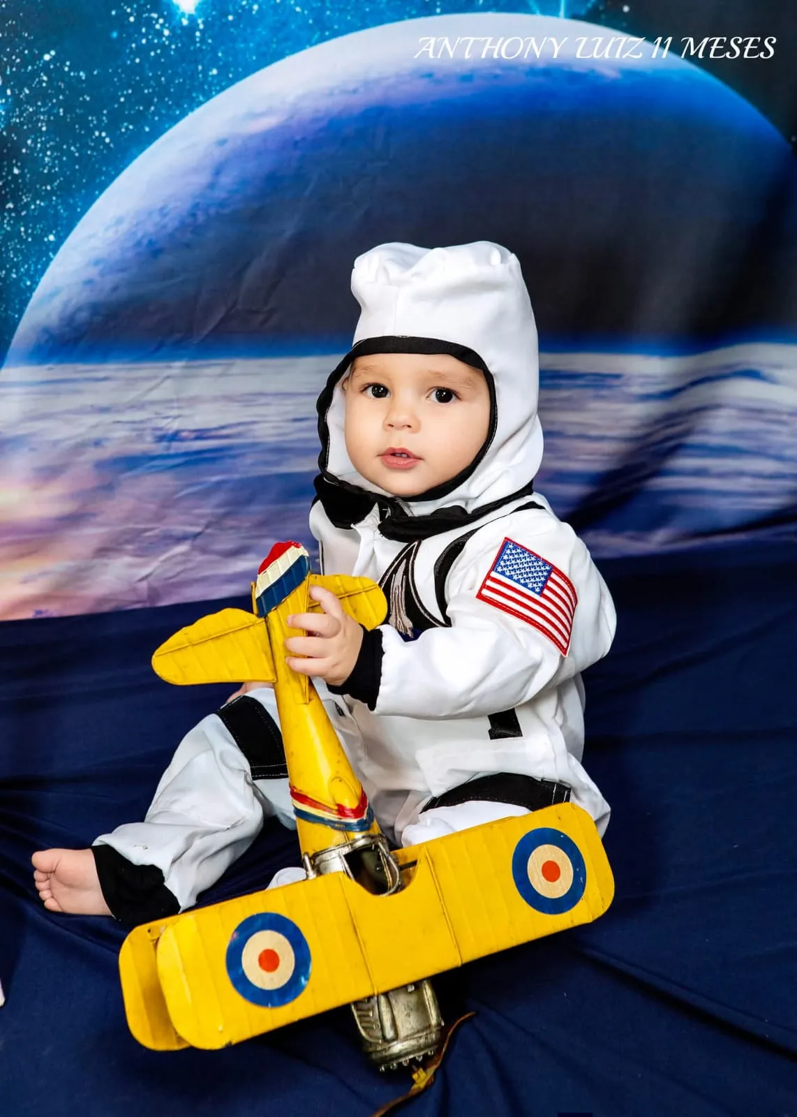 Bebê de 11 meses fantasiado de astronauta sobre avião de brinquedo, em cenário espacial.