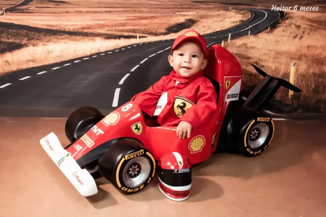 Bebê de 6 meses de macacão vermelho em carrinho de corrida, em cenário de estrada.