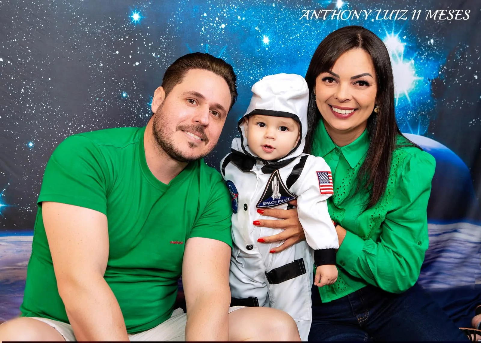 Bebê de 11 meses fantasiado de astronauta no colo dos pais, em cenário espacial.