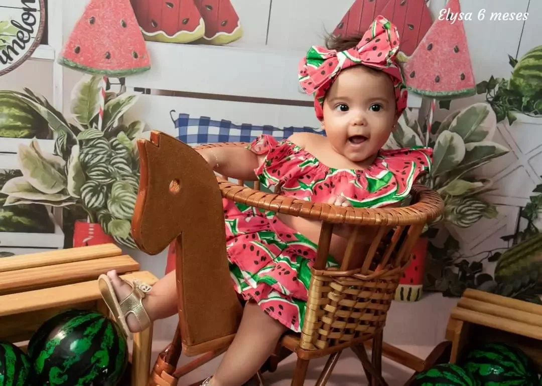 Bebê de 6 meses de vestido estampado de melancias sentada em cadeira de balanço de madeira.