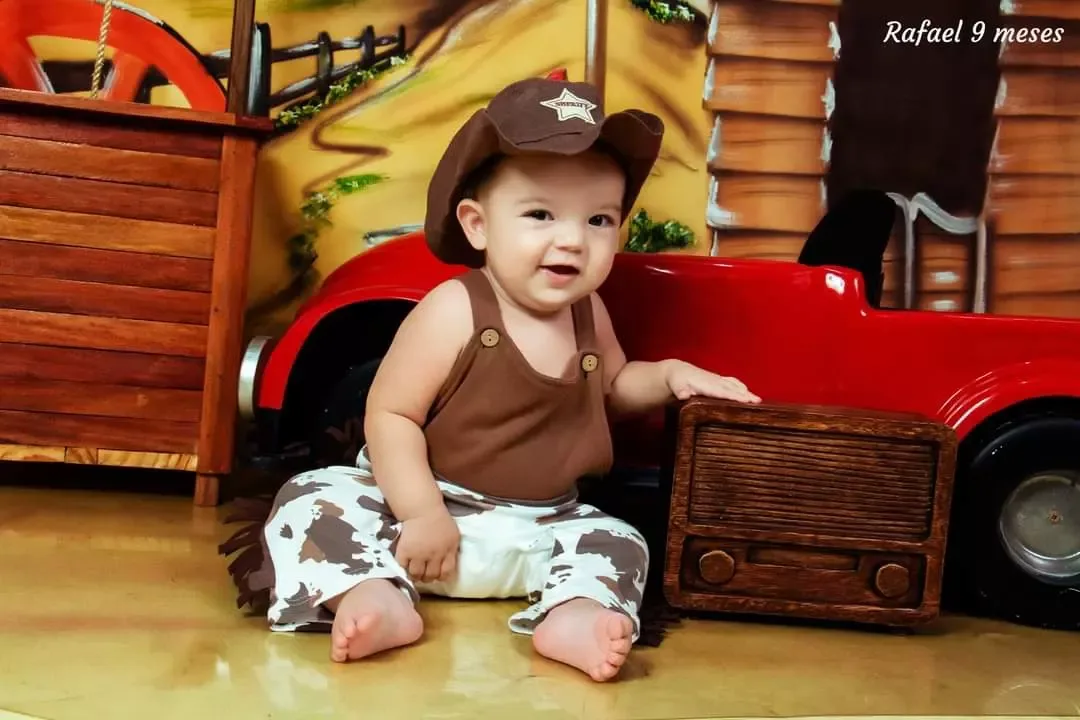 Bebê de 9 meses de chapéu de vaqueiro diante de caminhonete vermelha de brinquedo.