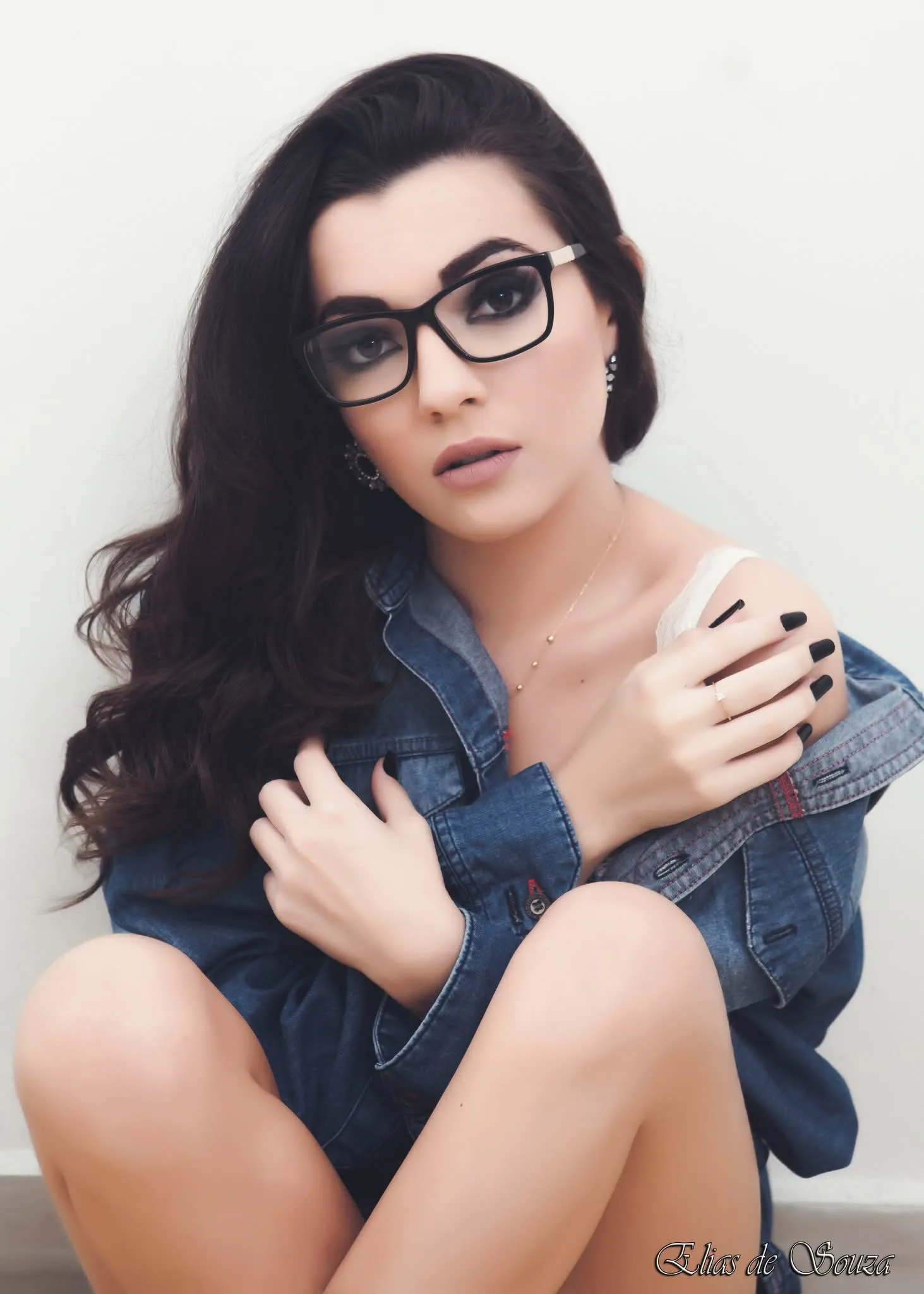 Mulher de óculos e jaqueta jeans sentada em fundo claro.