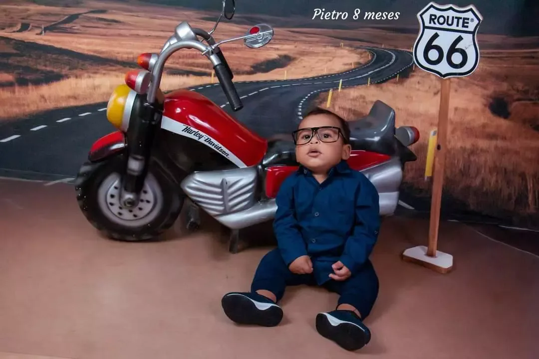 Bebê de 8 meses de óculos e camisa azul sentado ao lado de moto de brinquedo.