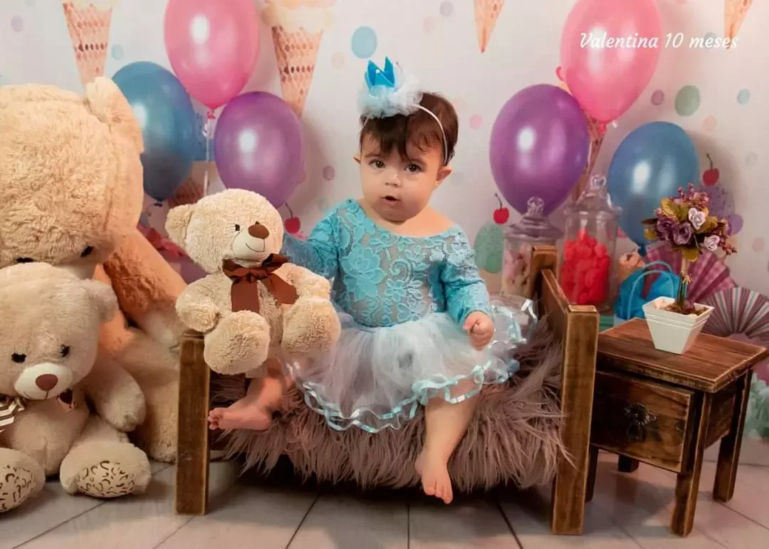 Bebê de 10 meses de vestido azul entre ursos de pelúcia e balões.