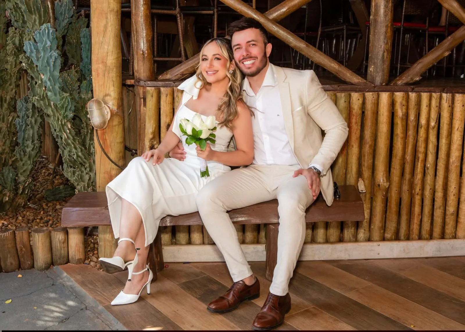 Casal de roupas claras sentado em banco de madeira, noiva com buquê branco.