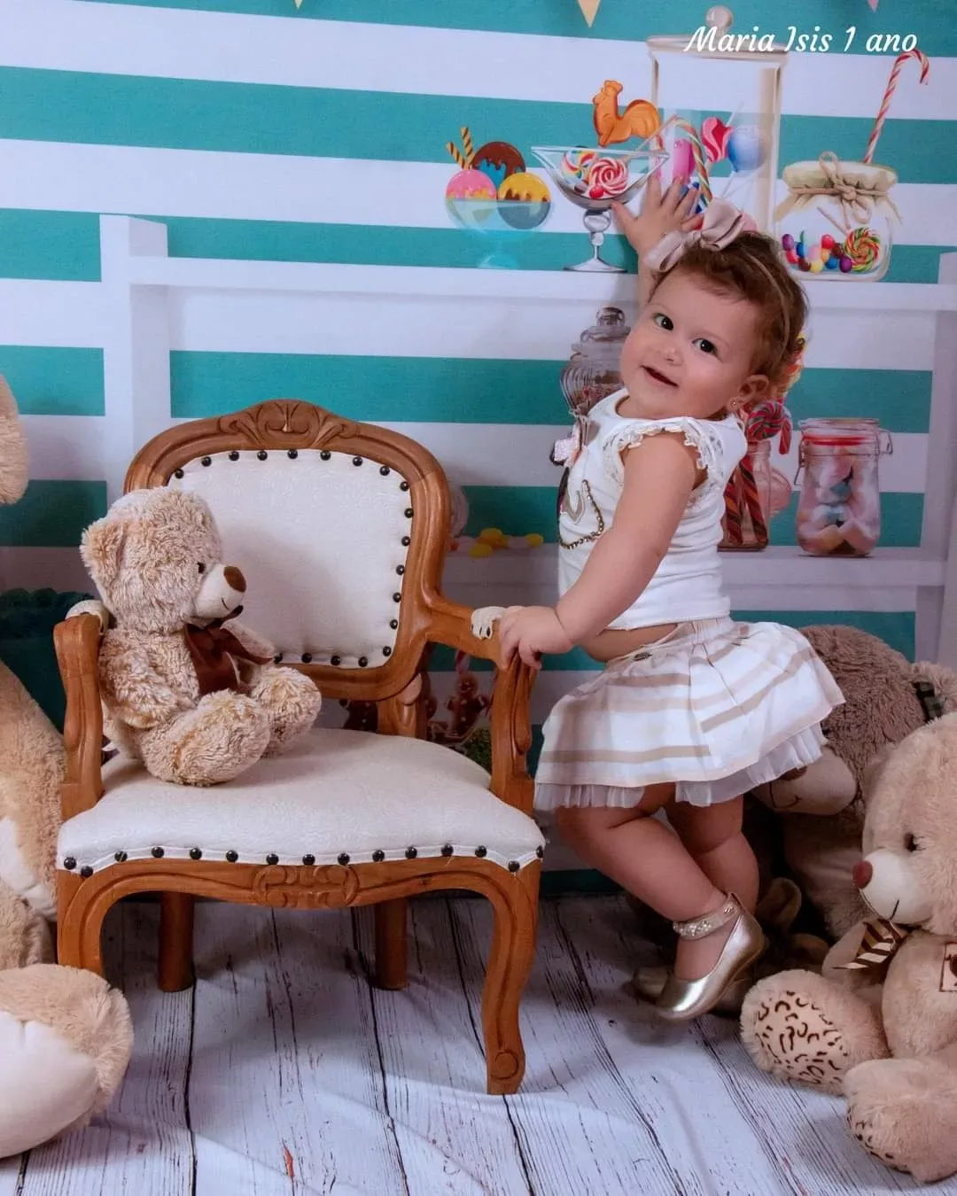 Menina de 1 ano em pé ao lado de poltrona e urso de pelúcia.
