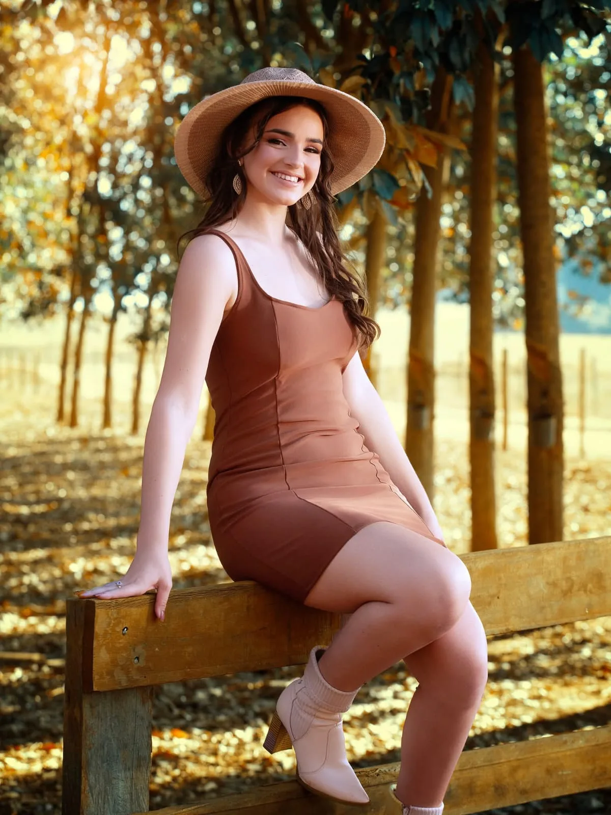 Mulher de chapéu e vestido marrom sentada em cerca de madeira.