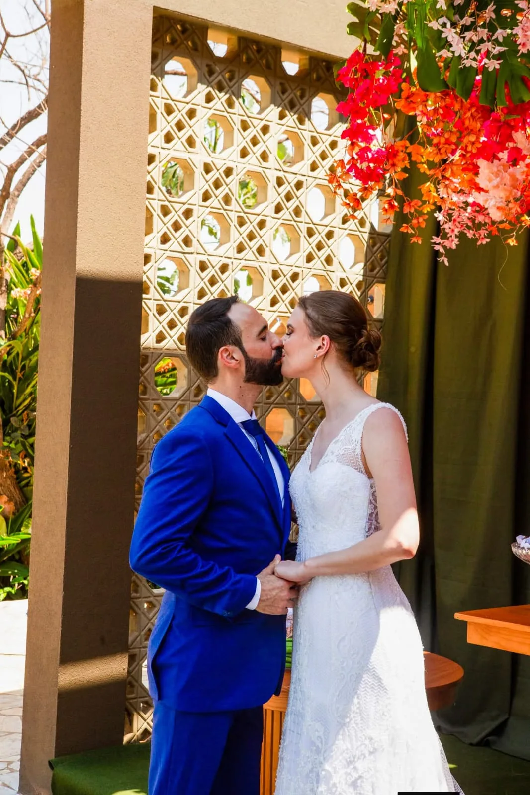 Noivos se beijando diante de portal de madeira com flores vermelhas.