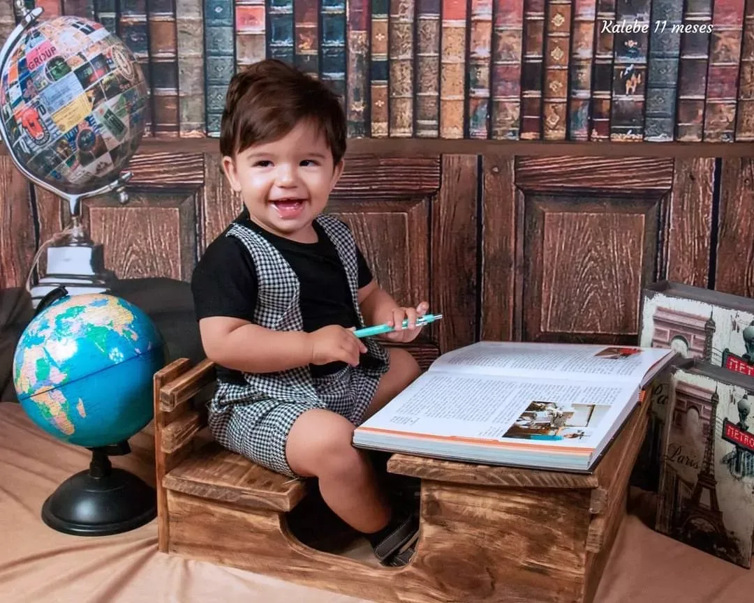 Bebê de 11 meses de suspensórios sentado em carteira escolar diante de livros.
