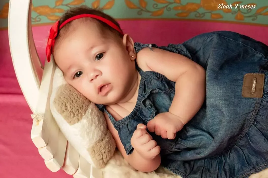 Bebê de 3 meses de jardineira jeans e faixa vermelha deitada em almofada.
