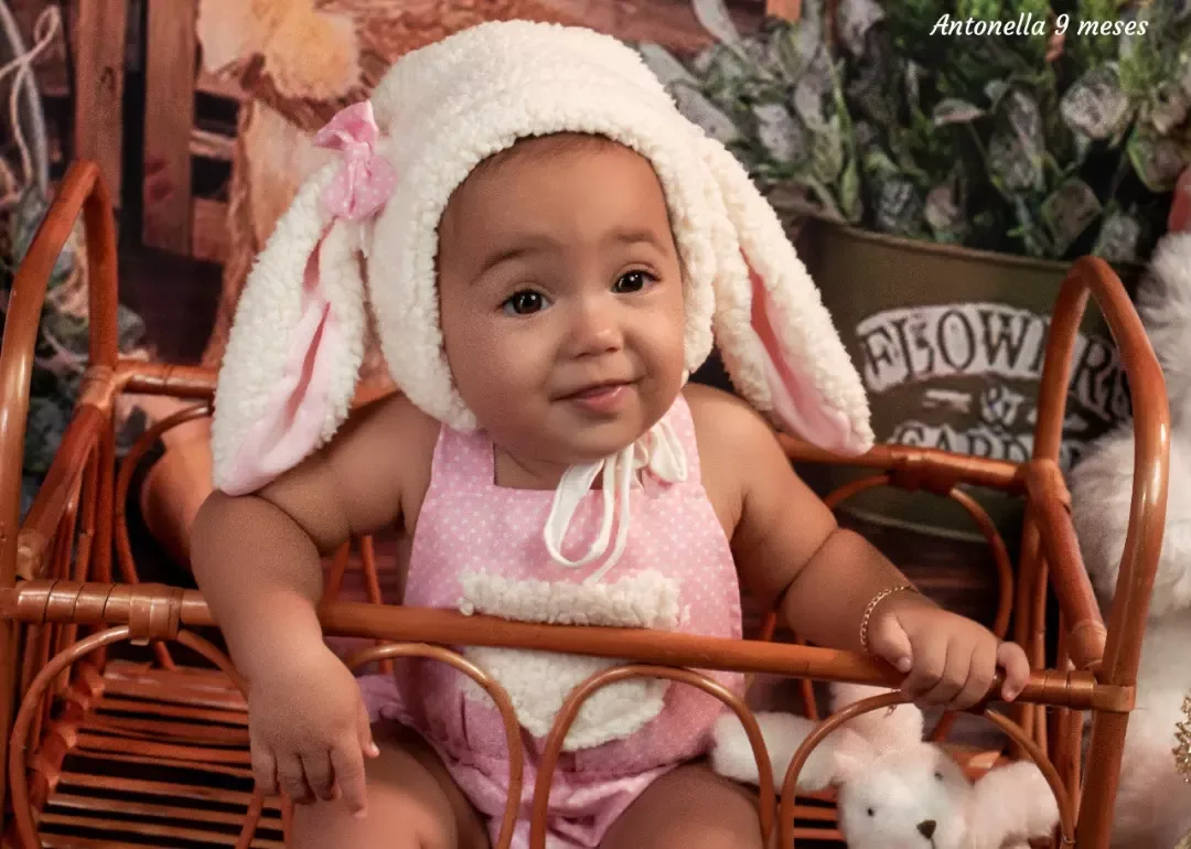 Bebê de 9 meses com touca de coelho sentada em poltrona de vime.