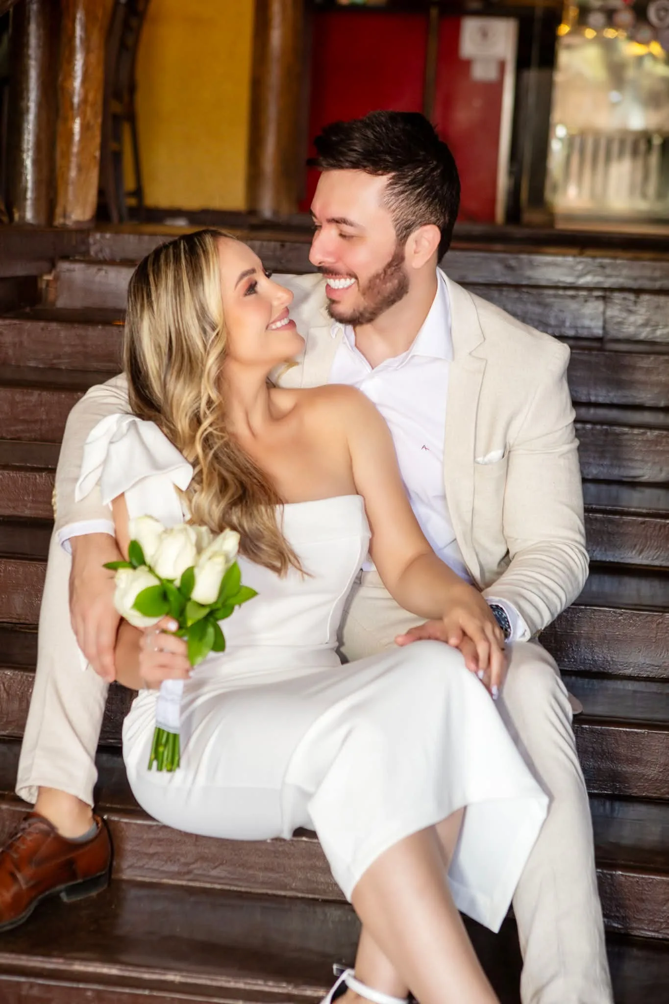 Casal de roupas claras sentado em banco de madeira, sorrindo um para o outro.