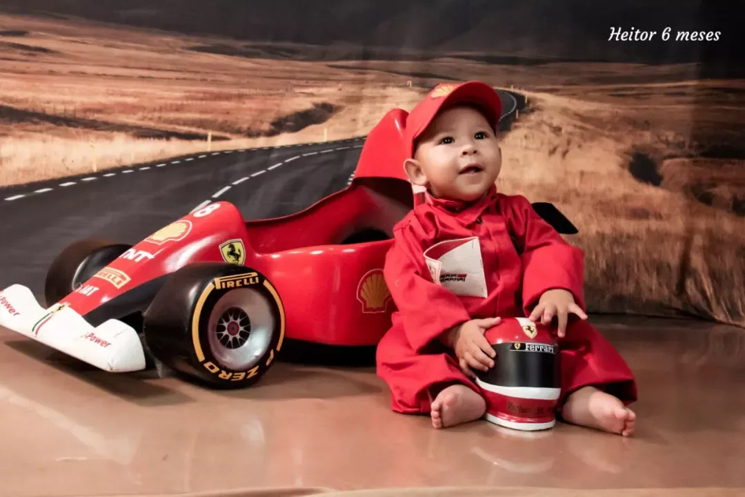 Bebê de 6 meses de macacão vermelho sentado ao lado de carrinho de corrida.