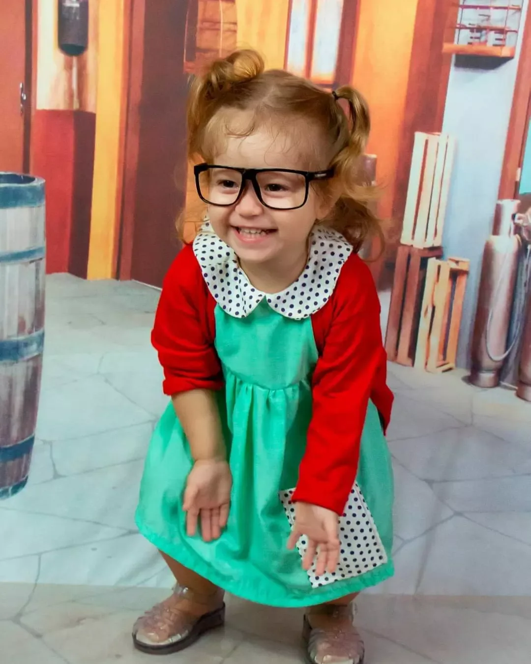 Menina de óculos, casaco vermelho e vestido verde sorrindo.