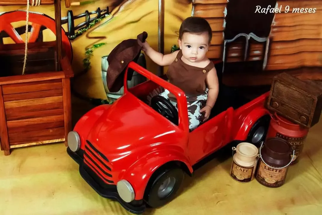 Bebê de 9 meses dentro de caminhonete vermelha de brinquedo em cenário de fazenda.