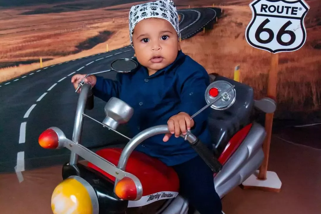 Bebê de 8 meses de chapéu sobre moto de brinquedo, com placa Route 66 ao fundo.