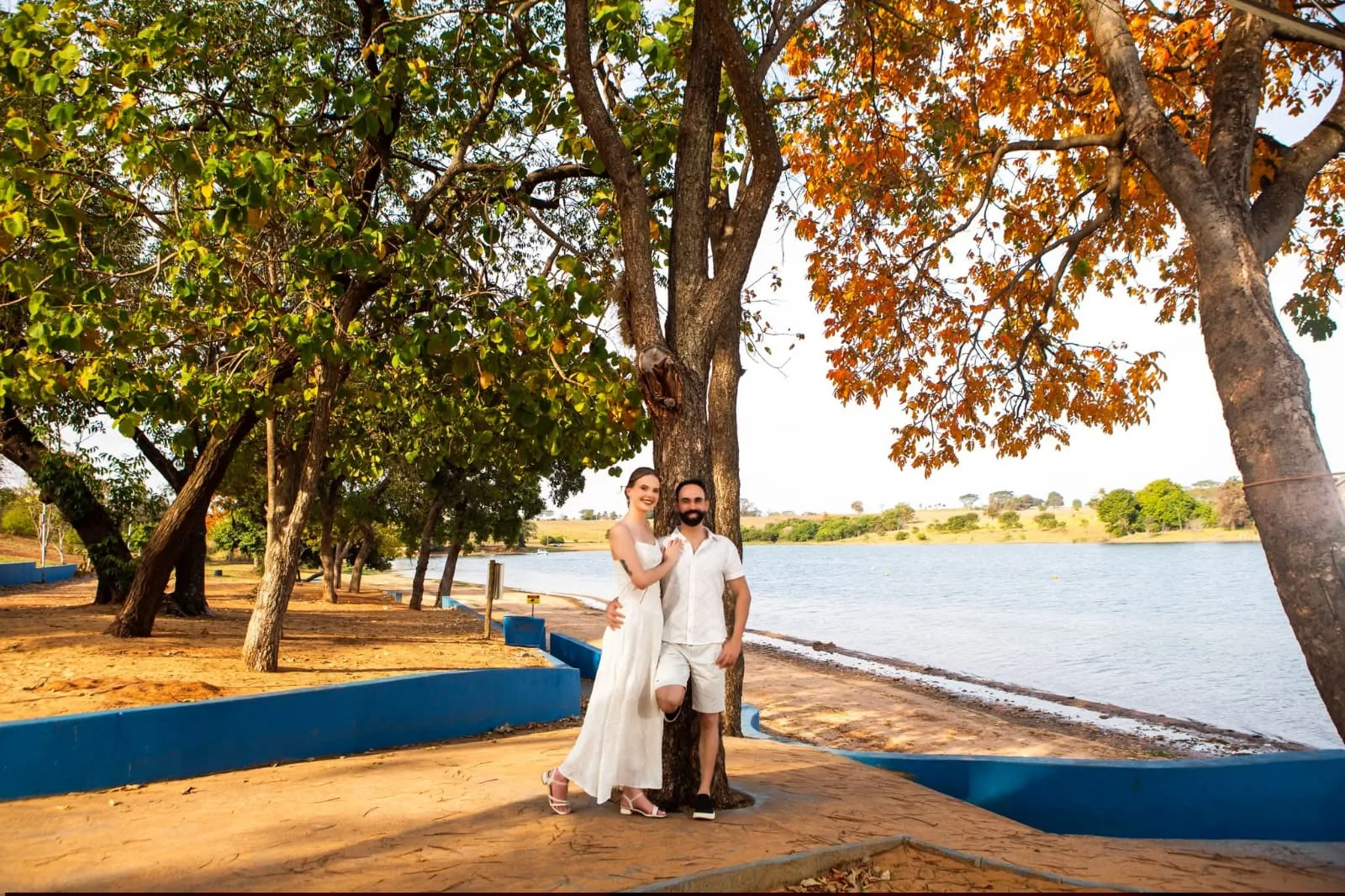 Casal de roupas claras à beira de um lago, entre árvores.