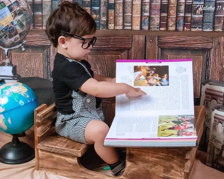 Bebê de 11 meses de óculos folheando um livro em cenário de biblioteca.