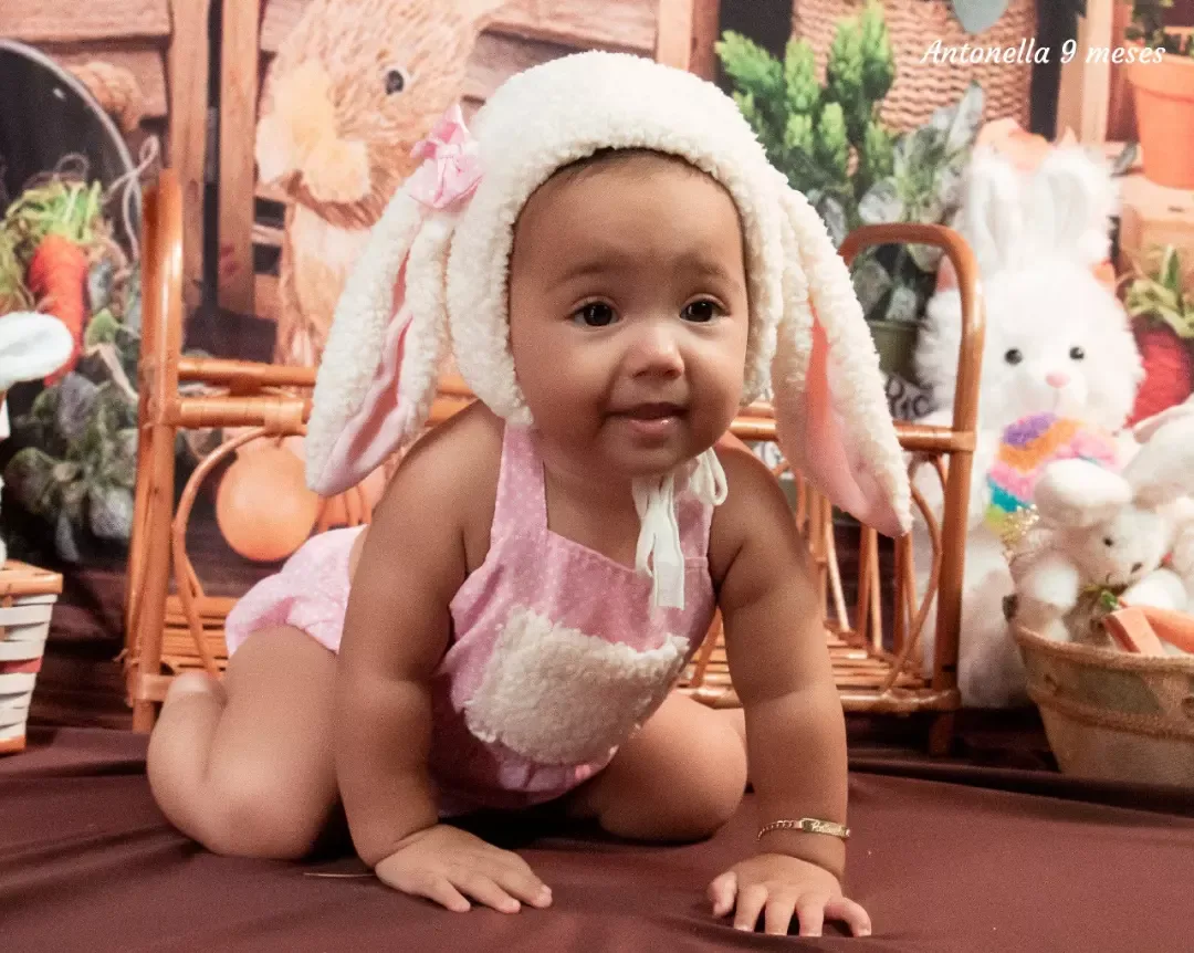 Bebê de 9 meses com touca de coelho engatinhando em cenário de Páscoa.
