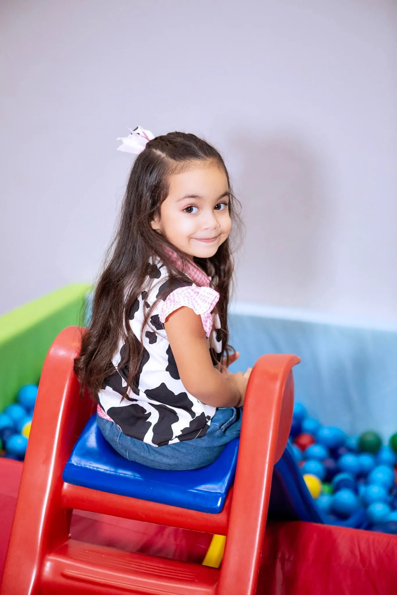 Menina de roupa estampada de vaca sentada em escorregador, junto à piscina de bolinhas.