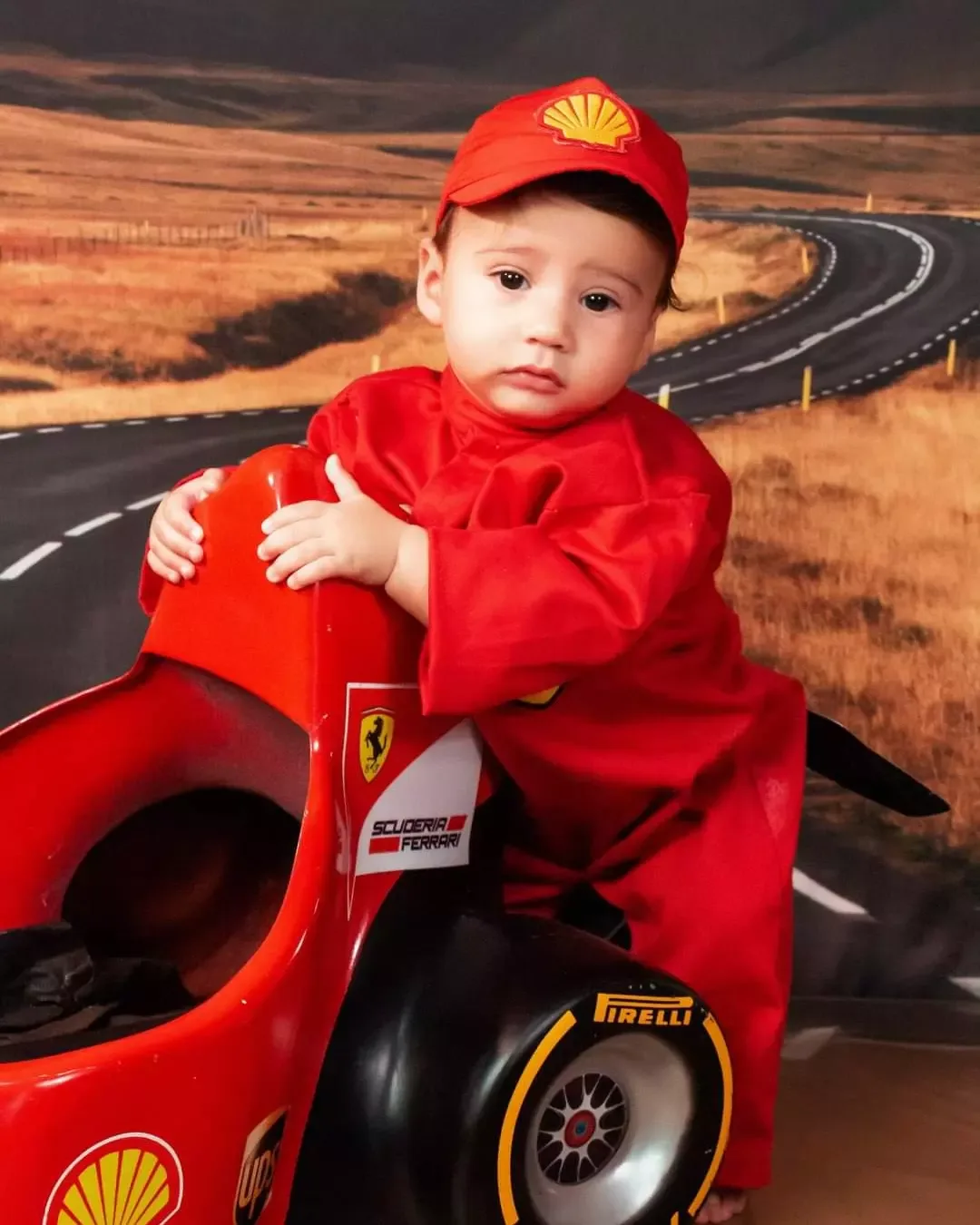 Bebê de 6 meses de macacão e boné vermelhos apoiado em carrinho de corrida.
