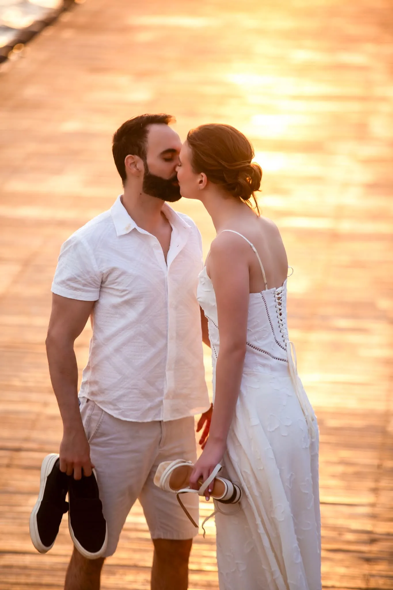 Casal se beijando ao pôr do sol, noiva segurando os sapatos.
