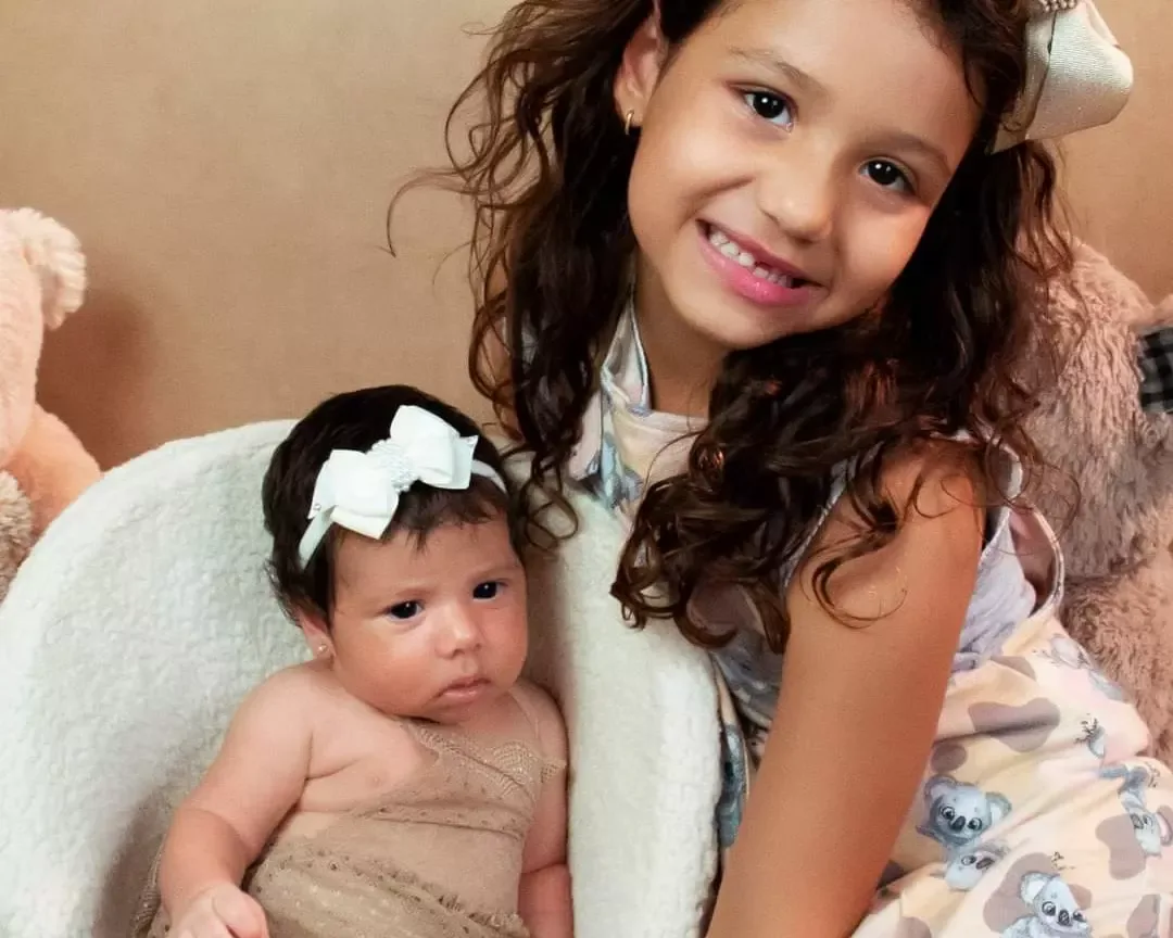 Recém-nascida de 1 mês com laço branco ao lado de uma menina sorrindo.
