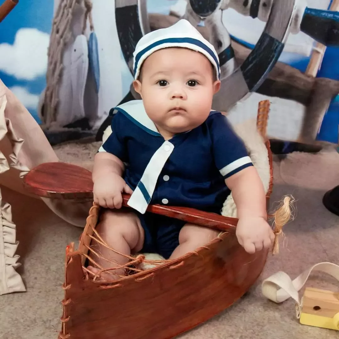 Bebê de roupa de marinheiro sentado em barquinho de madeira.