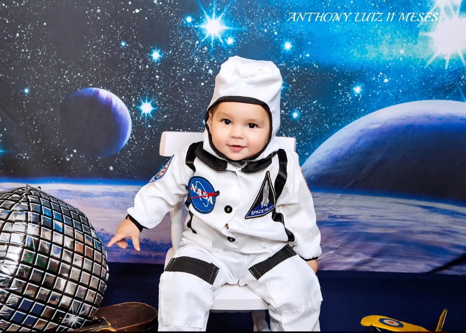 Bebê de 11 meses de roupa de astronauta sentado em cenário espacial com globo espelhado.